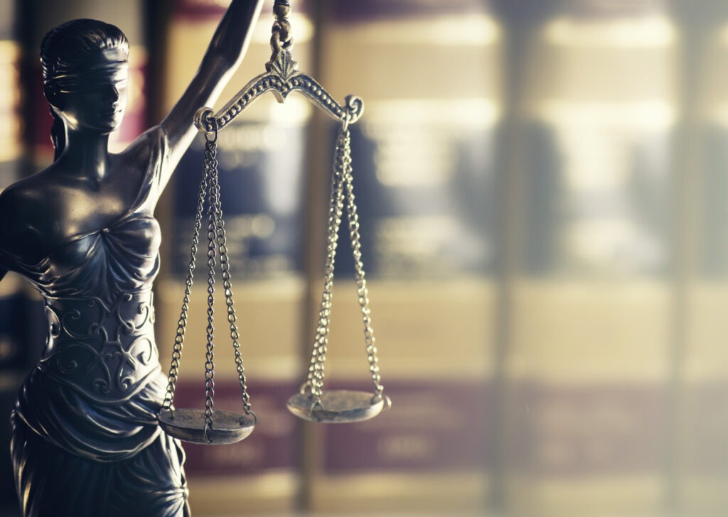 The Scales of Justice held by a blindfolded statue represent impartiality in law, set against blurred legal books, emphasizing justice's foundation.