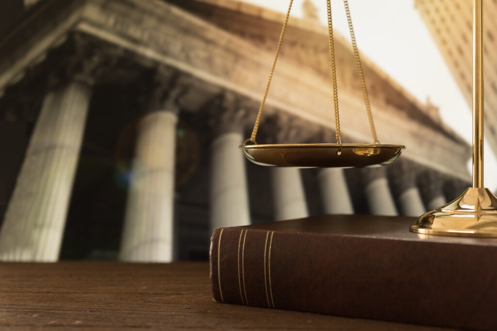 Golden scales of justice above a brown law book, with classic columns blurred in the background, symbolizing the legal system and justice.