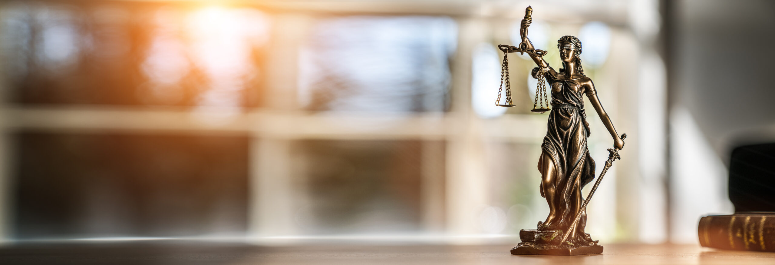 Bronze statue of Lady Justice holding scales and a sword, symbolizing law and fairness, with soft lighting in a legal setting.